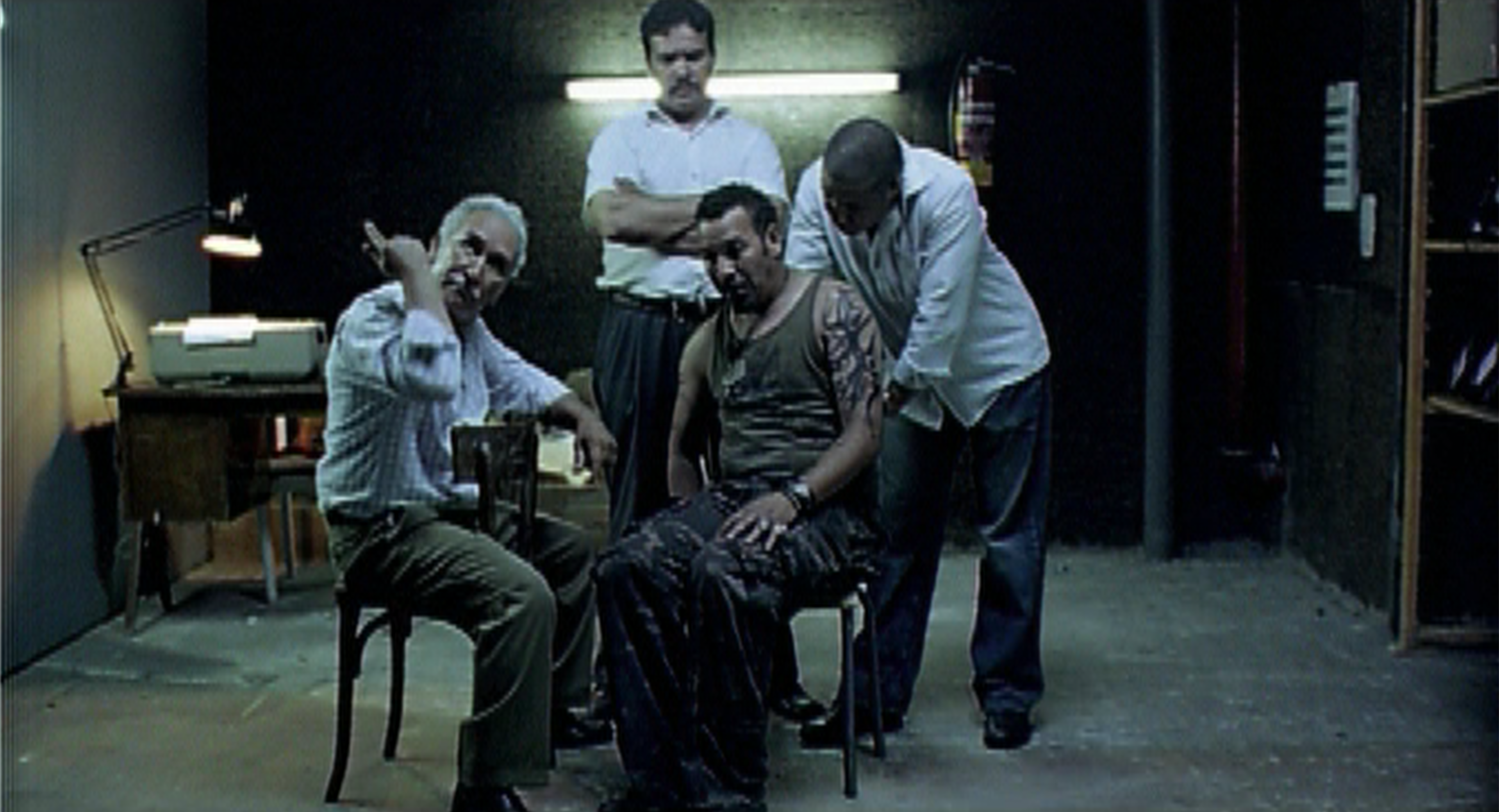 Dans un local sombre éclairé par un néon, un homme en débardeur assis sur une chaise, encadré par trois hommes&nbsp;; l’un d’eux pointe le doigt vers lui.