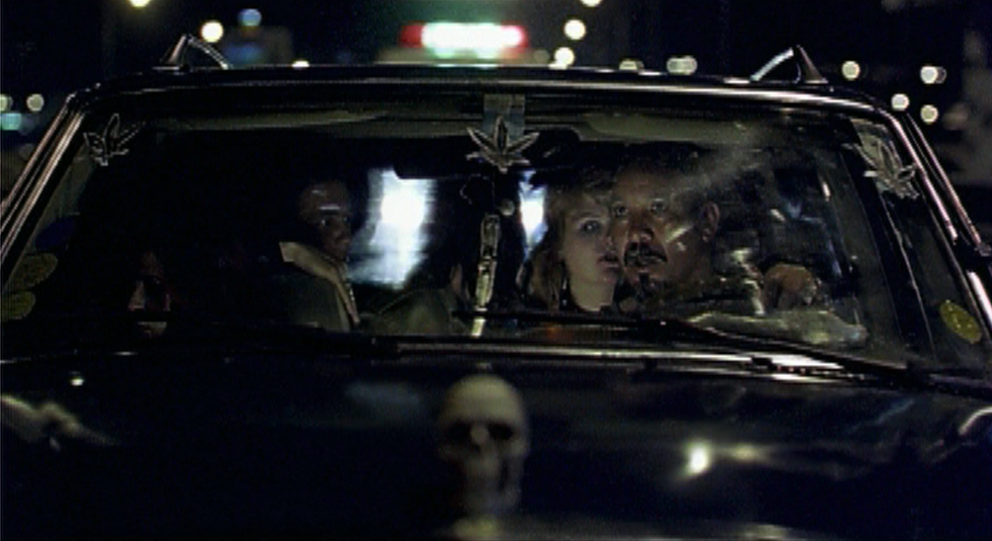 De nuit, à travers le pare-brise d’une voiture, un homme et une femme assis à l’avant&nbsp;; une tête de mort décore le capot.