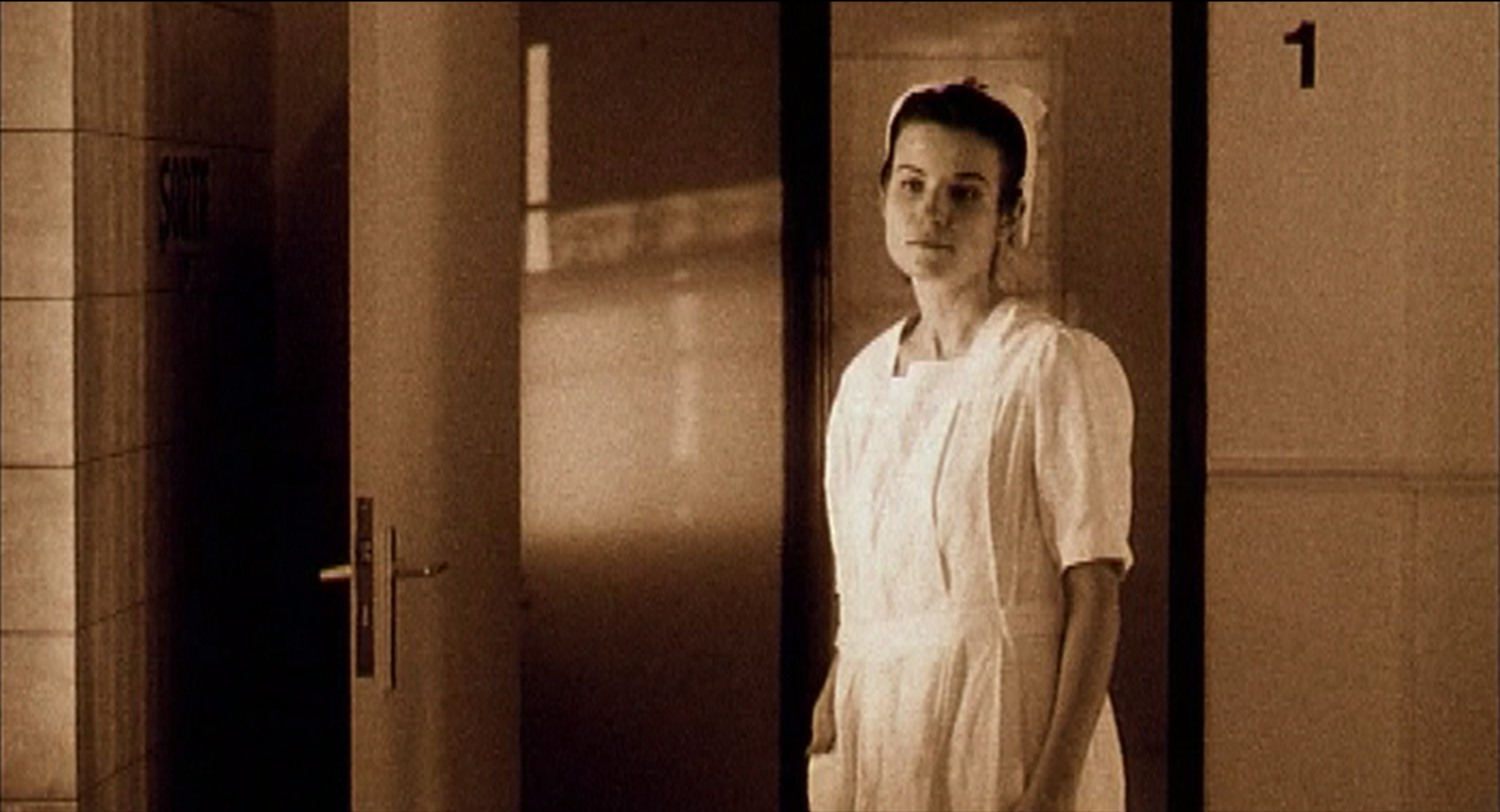 Une jeune femme en blouse et coiffe blanches se tient dans l’embrasure d’une porte, au fond d’un couloir carrelé.