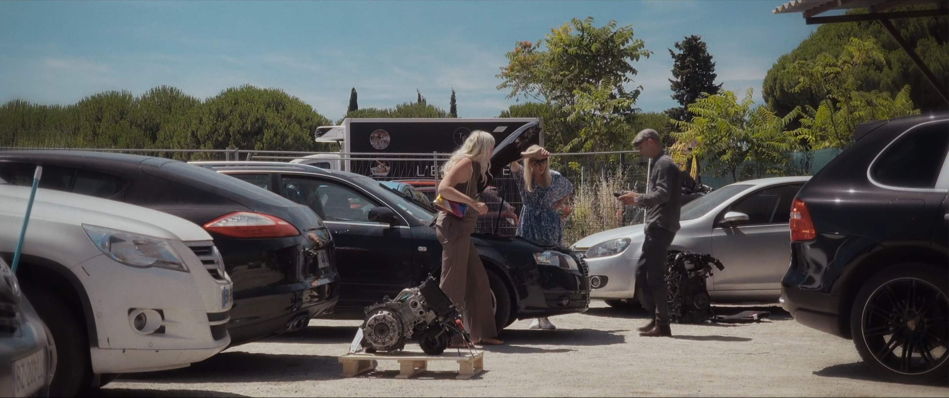 Sur un parking en plein soleil, deux femmes et un homme debout entre des voitures, près de pièces mécaniques posées au sol.