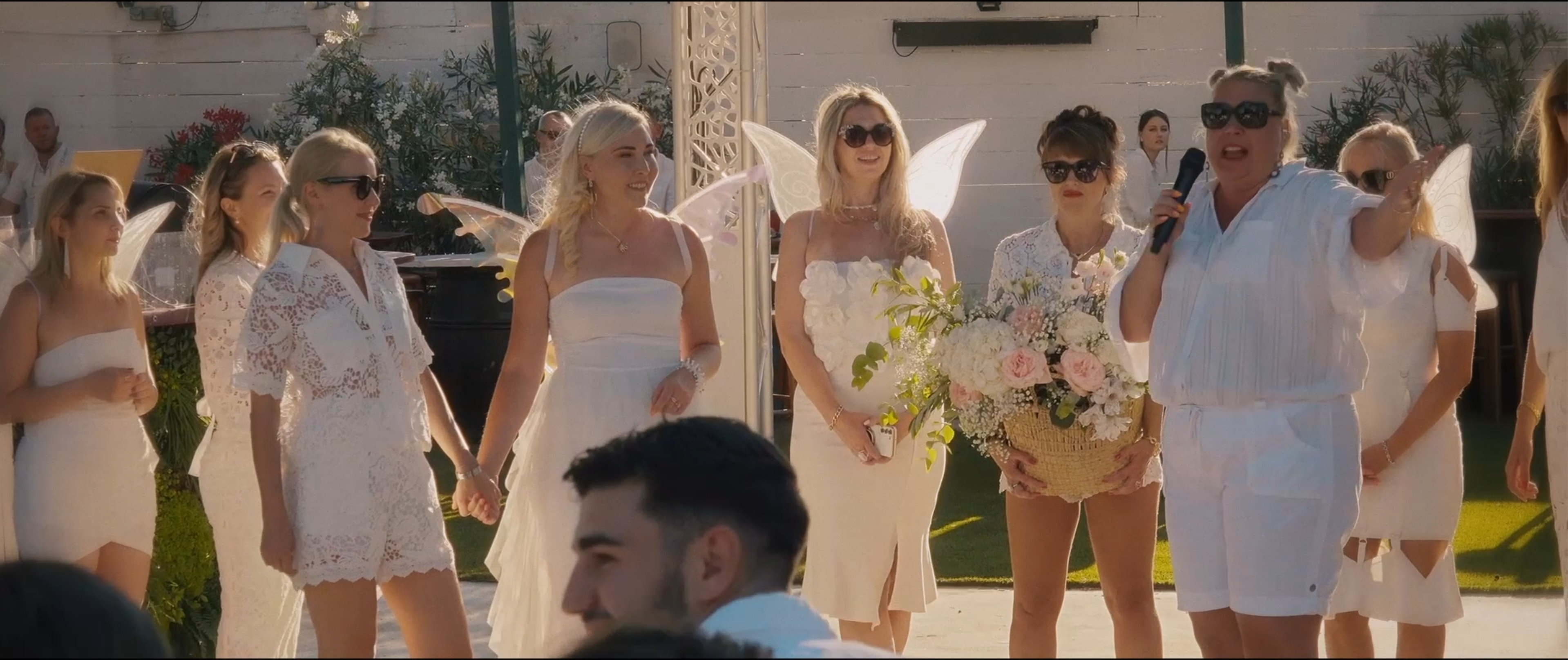 Lors d’une fête en plein air, des femmes en blanc, certaines avec des ailes d’ange, entourent l’une d’elles qui parle au micro&nbsp;; une autre tient un gros bouquet.